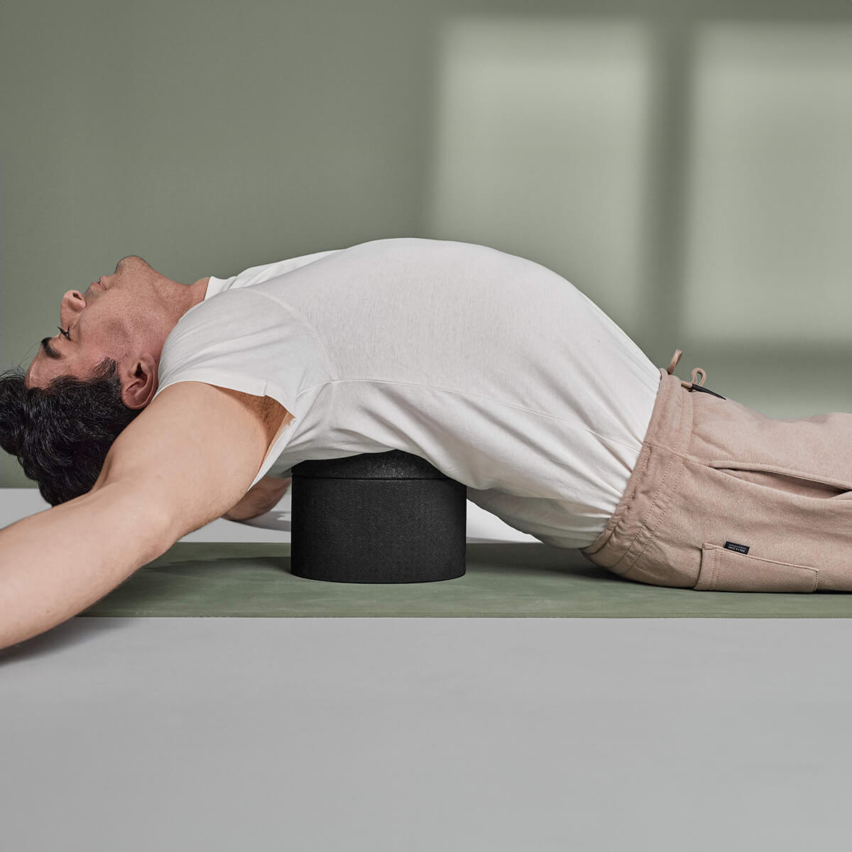 Eine Person in weißem Hemd und beiger Hose liegt auf einer Yogamatte und benutzt die Liebscher & Bracht Rückenretter Schaumstoffrolle unter dem oberen Rücken, um diesen zu dehnen und die Haltung zu verbessern - ideal zur Linderung von Rückenschmerzen.